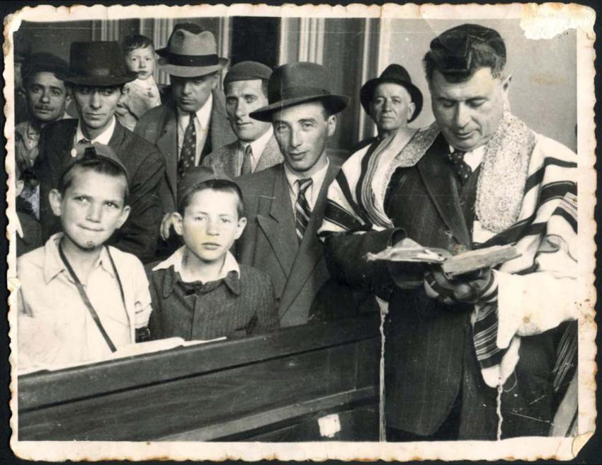 חזן וילדים עוטי טלית ותפילין במהלך חגיגת בר מצווה בבית יתומים, במרכז התצלום נראה מנהל המוסד מנחם גלזר, בוקרשט, רומניה, 1944