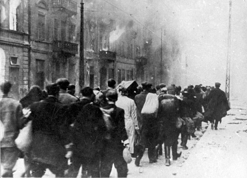 Jews being led to the Umschlagplatz (the deportation area) during the time of the uprising. In the background, the ghetto buildings can be seen going up in flames