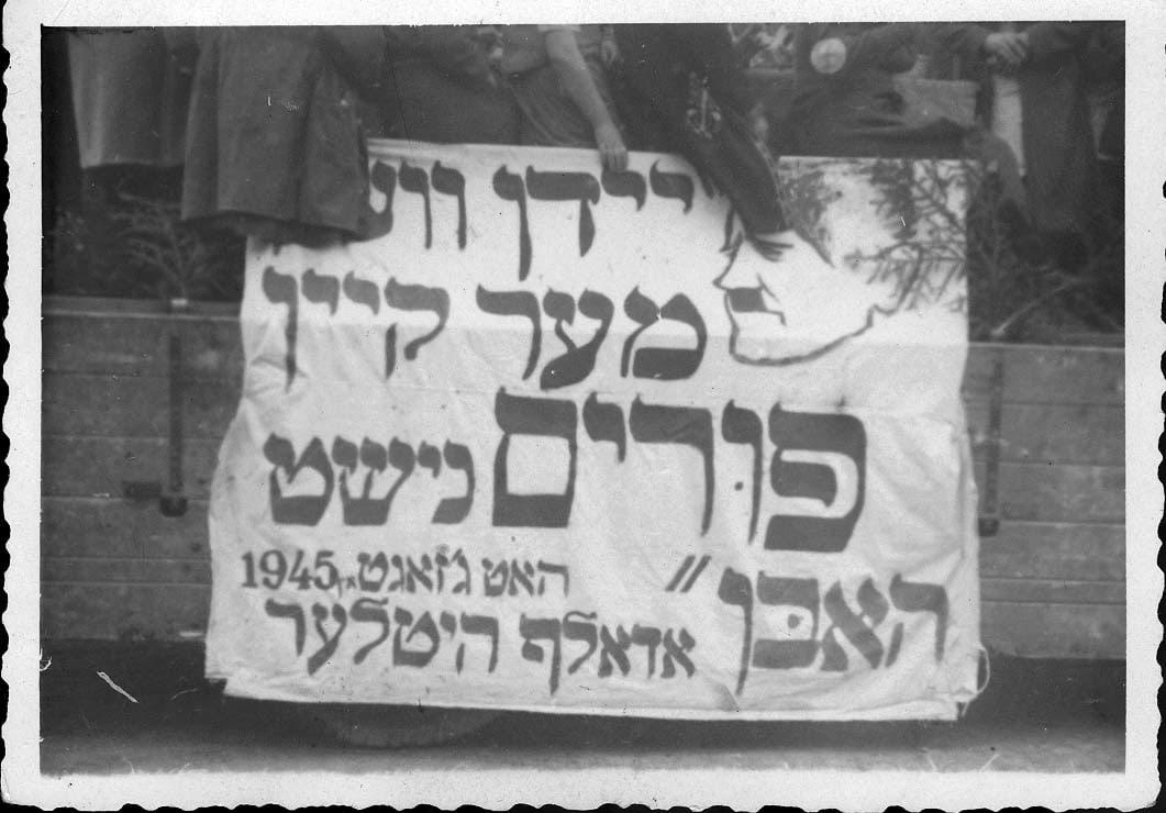 שלט עם ציטוט המיוחס להיטלר, "יהודים כבר לא יחגגו פורים", מחנה העקורים לנדסברג, 1946 בערך