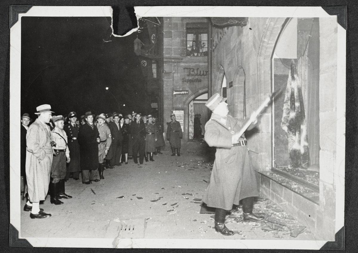 A crowd of bystanders including civilians, SS and SA men looking at an SS member shattering a Jewish owned shop during the Kristallnacht, Nürnberg, 1938. Bystanders watching the destruction of a Jewish shop in Nürnberg.