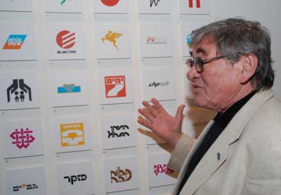 חתן פרס ישראל לעיצוב, ניצול השואה דן רייזינגר, על רקע סמלים שונים בעיצובו המוצגים בתערוכה