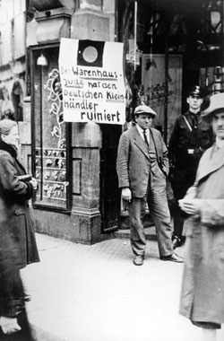 Würzburg, „Tag des Boykotts“, 1. April 1933. Ein SS-Mann steht vor einem jüdischen Geschäft. Auf dem Schild steht: „Der Warenhausjude hat den deutschen Kleinhändler ruiniert“