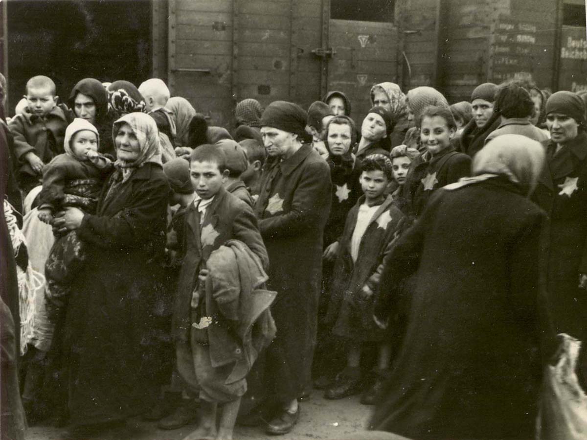 Die angekommenen Juden erwarten vor den Güterwaggons die Befehle der Deutschen. Einige bemerken den Fotografen und blicken neugierig in die Kamera