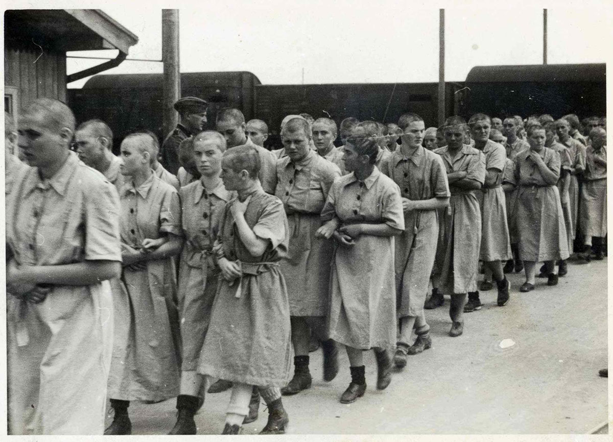 Jewish women prisoners walk inside the women's camp, wearing the standard prison uniform. On the left, a SS man watches them