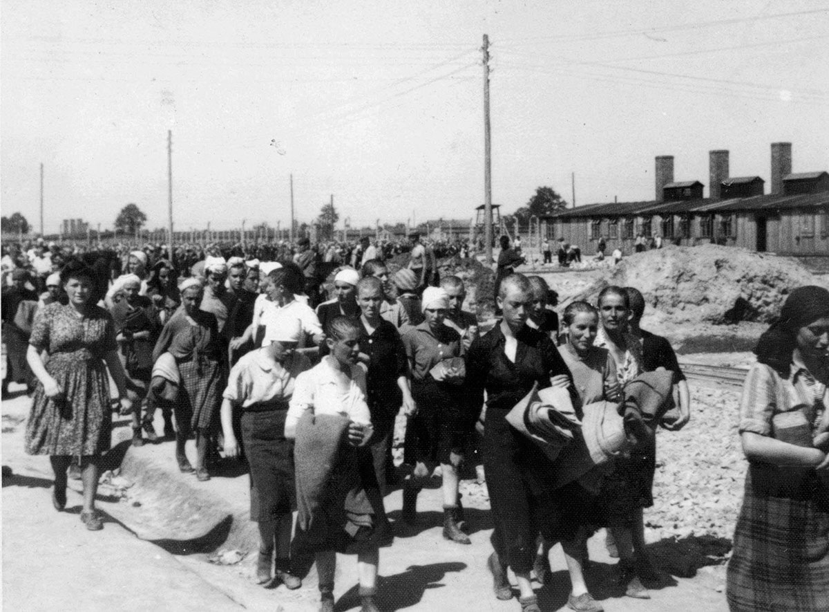 Prisioneras siendo llevadas a las barracas por la Blockälteste (jefa de bloque, a la izq.); el edificio a la derecha es la cocina del campo para mujeres