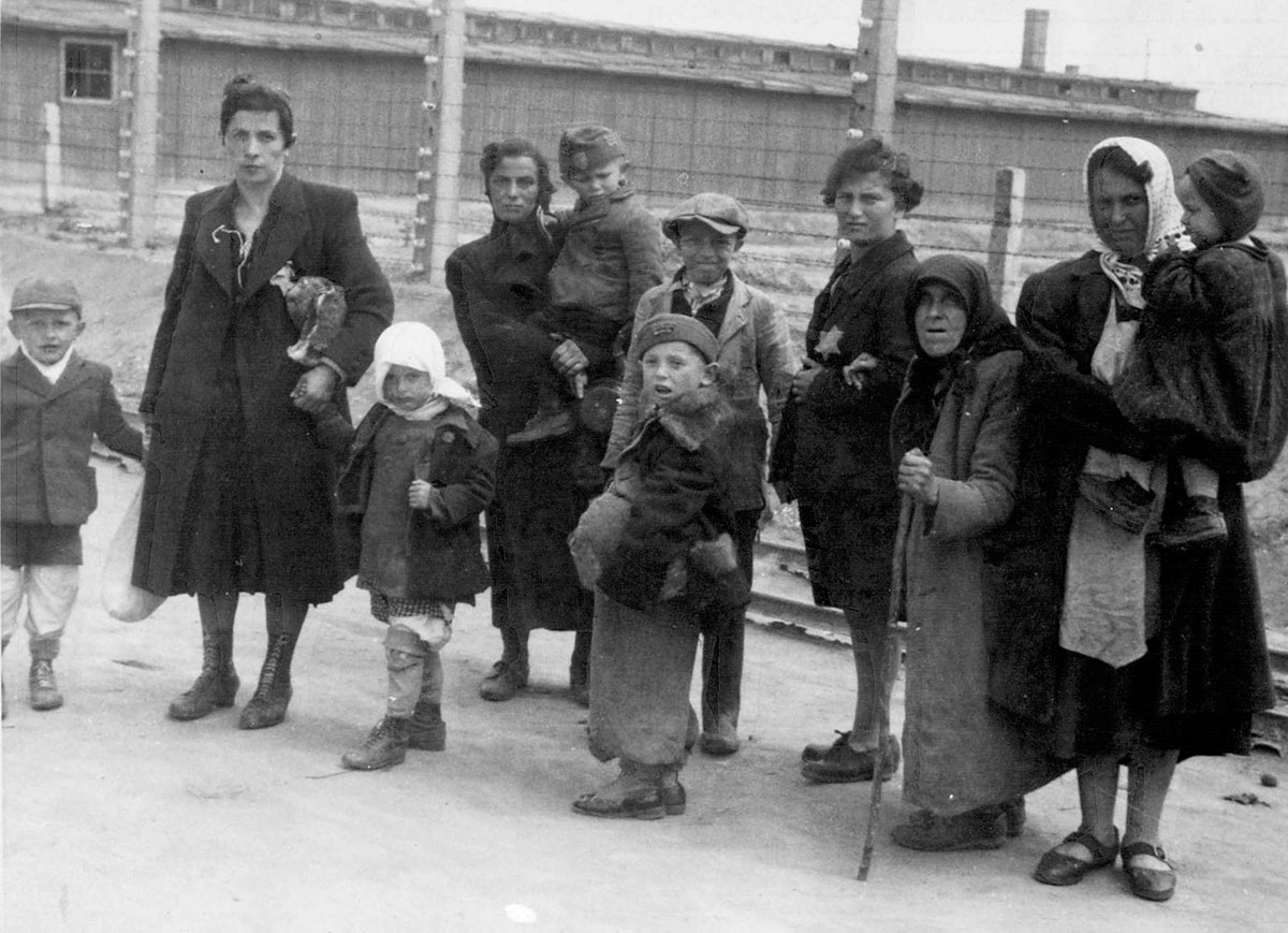 Mères et enfants juifs forcés à aller vers les chambres à gaz passant devant les baraques et la clôture électrifiée.