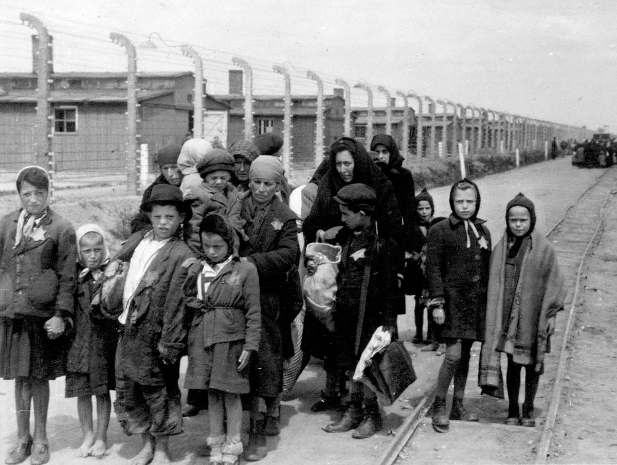 אמהות וילדים יהודיים המאולצים ללכת לתאי הגזים חולפים על פני הצריפים וגדר התיל המחושמלת