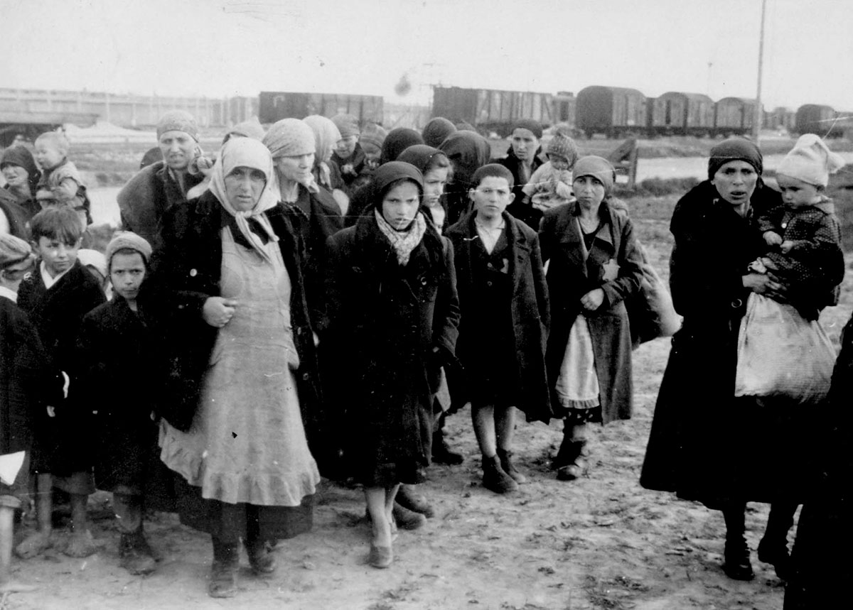 נשים יהודיות וילדים מאולצים ללכת אל תאי הגזים. ליד הרציף עדיין מחכים מספר קרונות רכבת