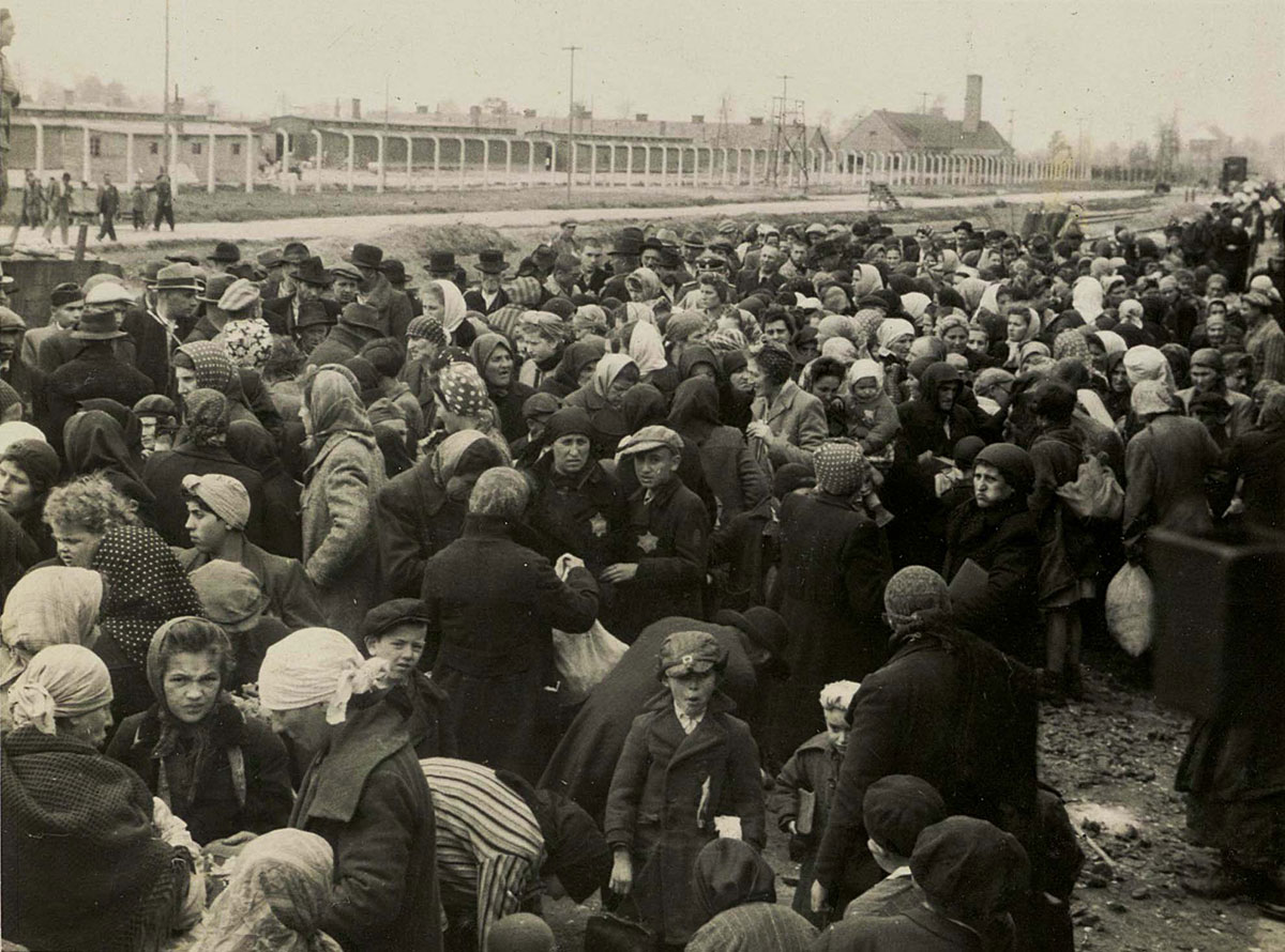 Certains Juifs regardent à l'intérieur du wagon, là où se trouve le photographe.  En haut à droite, le bâtiment du four crématoire II. En haut à gauche, « la rue du camp » (Lagerstrasse, appellation de la rue principale du camp)
