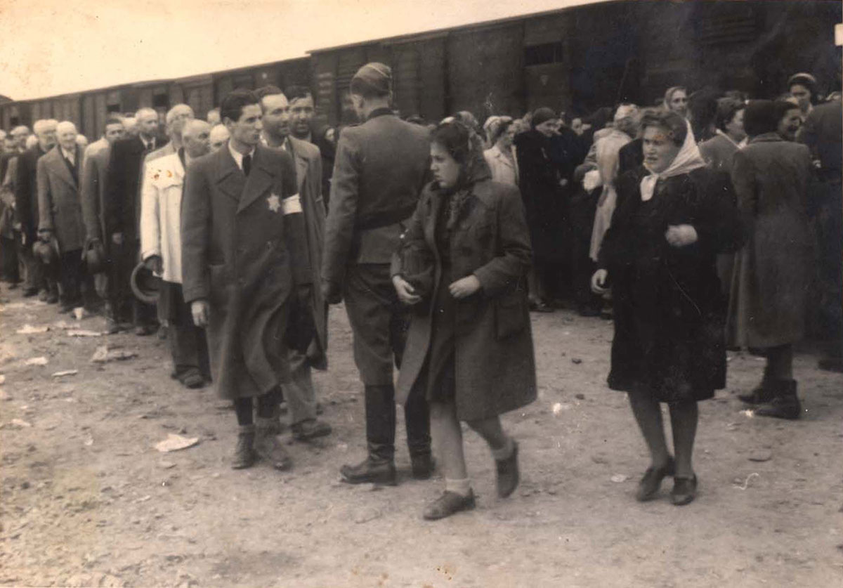 טור הגברים היהודים משמאל מתקדם לקראת איש אס-אס. ברקע - סלקציה של הנשים היהודיות. שני הנשים היהודיות בחזית נבחרו ככל הנראה לעבודת כפיה