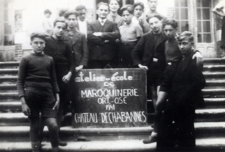 Leon König and his students with a sign that reads: "Leather Workshop-School, ORT-OSE, 1941, Château de Chabannes". On Leon König's right – Stephan Lewy, on Leon König's left – his son David