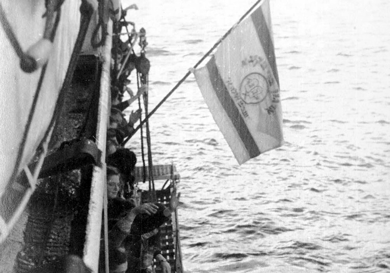 Members of Bnei Akiva from the "Ilania" children's village in Apeldoorn, the Netherlands waving their group's flag – Bnei Akiva, Shevet Ayala A – on board the "Negba" on their way to Israel.  Amsterdam, October 1948