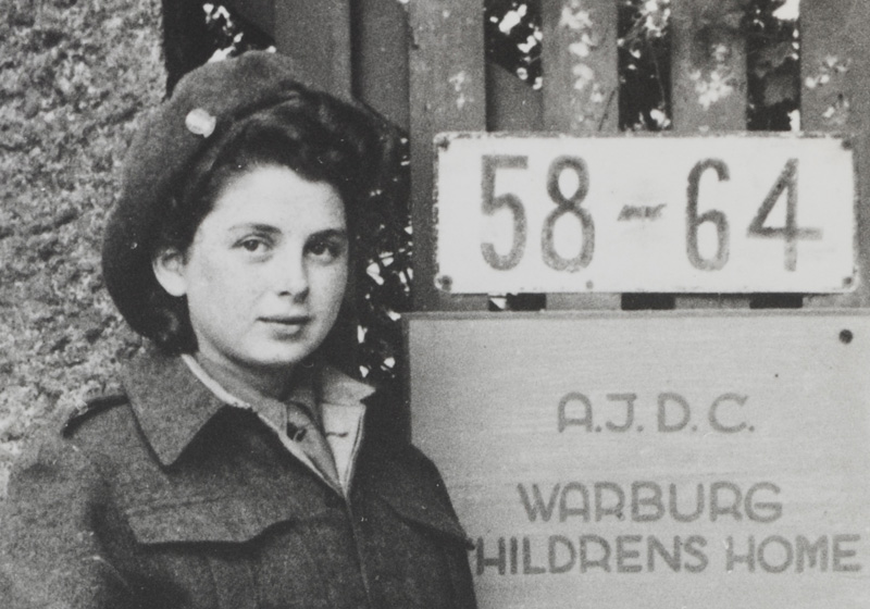 הנערה רניה באף (לימים רֶנֶה קוכמן) לצד שלט שער הכניסה לבית הילדים בבלנקנזה שבהמבורג, גרמניה, 1946-7